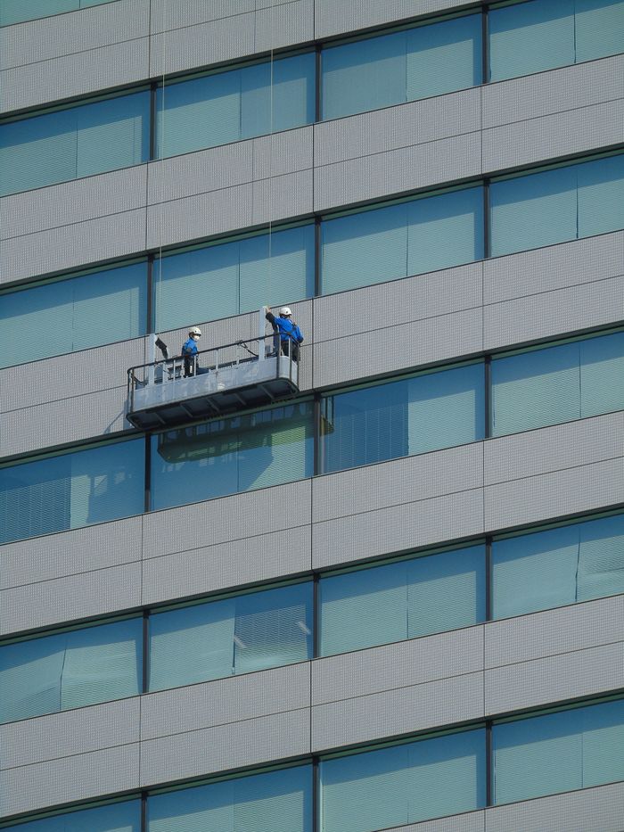 hero-img-01 Dos trabajadores limpian las ventanas de la fachada de un edificio moderno de vidrio utilizando una plataforma suspendida.